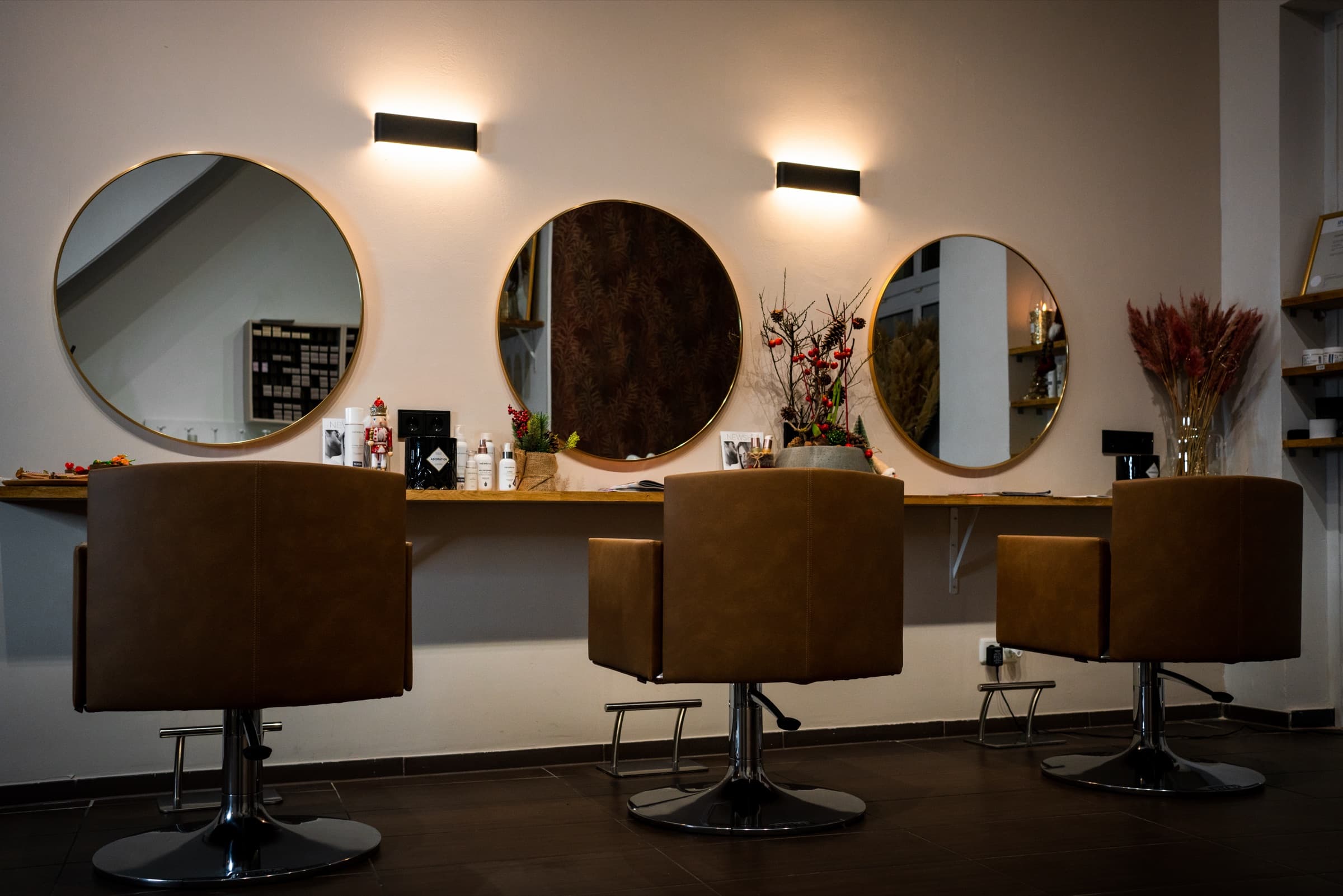 Drei Friseurplätze mit runden goldgerahmten Spiegeln, Wandleuchten und braunen Lederstühlen, dazwischen weihnachtliche Deko